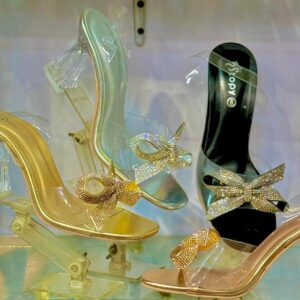 Display of four pairs of VOV crystal bow high heel sandals in a glass case: gold with rhinestone bow on clear platform heel, silver-clear version, black bow sandal, and nude-clear style.