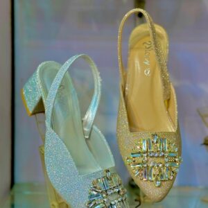 Pair of VOV glitter slingback block heel pumps displayed in a glass case: silver glitter pointed-toe shoe with large crystal brooch and 4-inch chunky heel on the left, gold glitter version on the right.