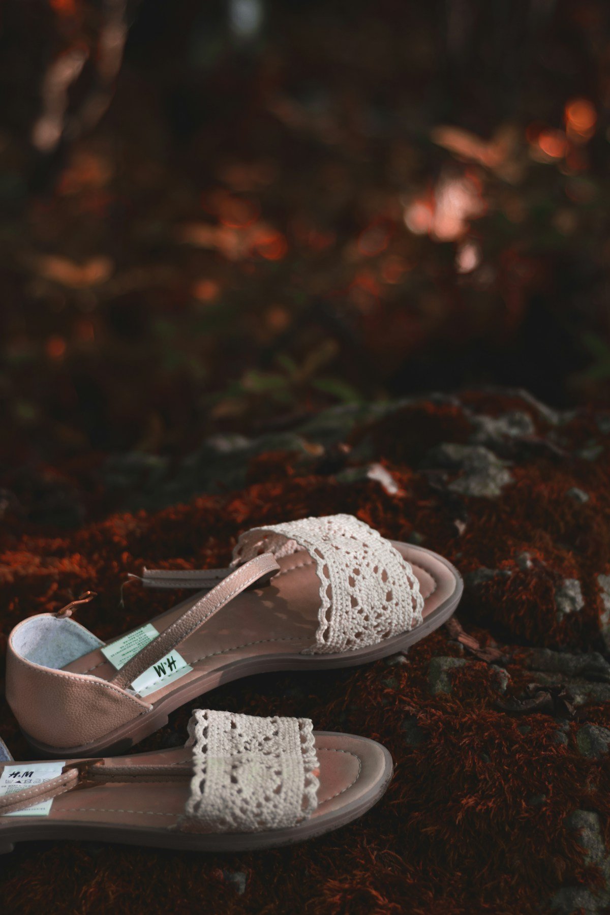 Light-coloured flat sandals with lace straps and closed heel sections on a woodland surface